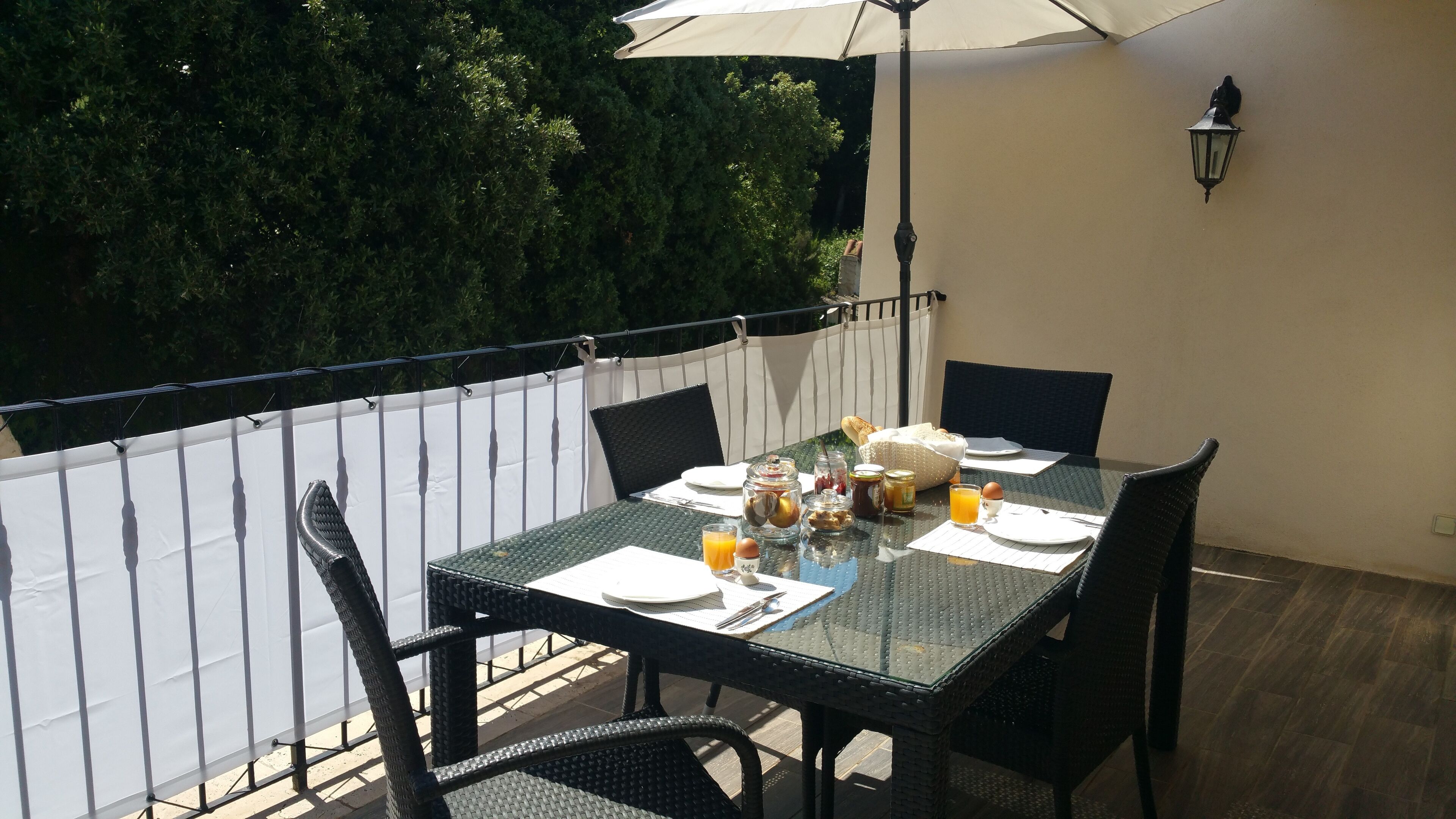 An outdoor dining area with a table set for two, surrounded by chairs, and an umbrella providing shade.