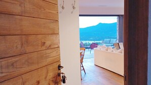 Una puerta de madera que da a una habitación con un sofá blanco, vistas a montañas y un balcón con sillas.