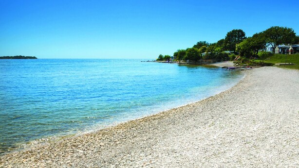 A pebble beach with clear blue water, a small island in the distance, and a grassy area with trees and a building. - Victoria Mobilehome Solaris fkk Resort (Tar-Vabriga)