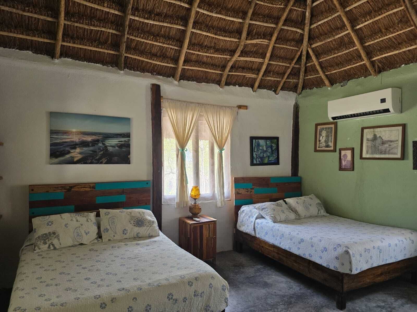 A room with two beds, a thatched roof, a window with curtains, and several framed pictures on the wall.