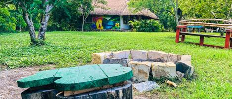 A rustic thatched-roof hut surrounded by lush greenery and a stone fire pit.
