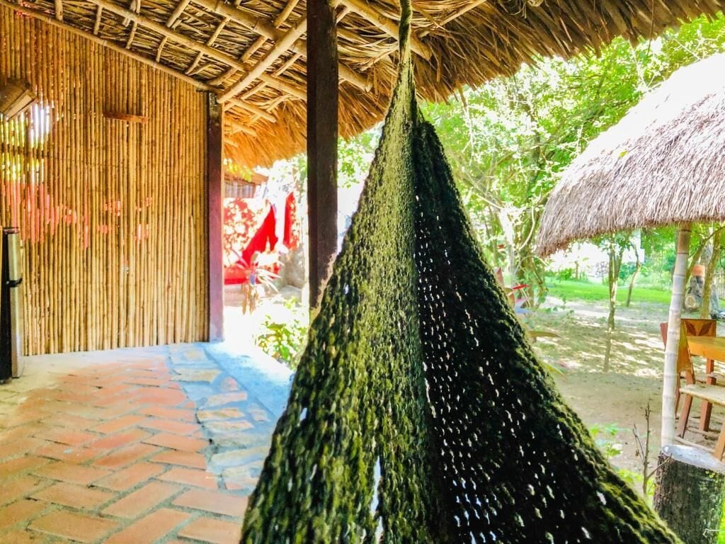 A hammock hanging from a bamboo structure with a thatched roof, a brick pathway, and a garden area with a thatched table and chairs.
