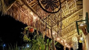 A thatched-roof outdoor area with a dreamcatcher hanging from the ceiling, surrounded by greenery.
