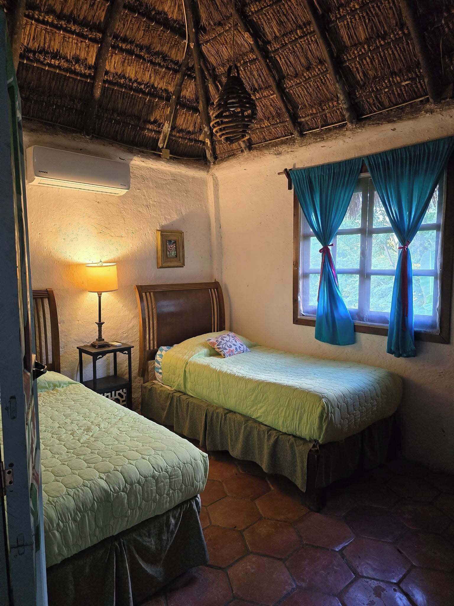 Una habitación rústica con techo de paja, dos camas individuales con ropa de cama verde, una mesita con una lámpara y una ventana con cortinas azules.