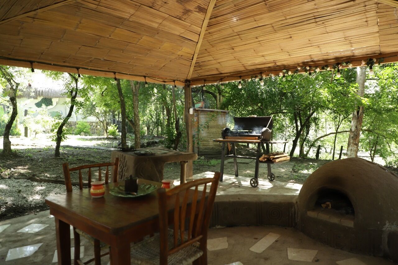 Un espacio para comer al aire libre con mesa y sillas, una parrilla y un horno de leña bajo un techo de bambú.