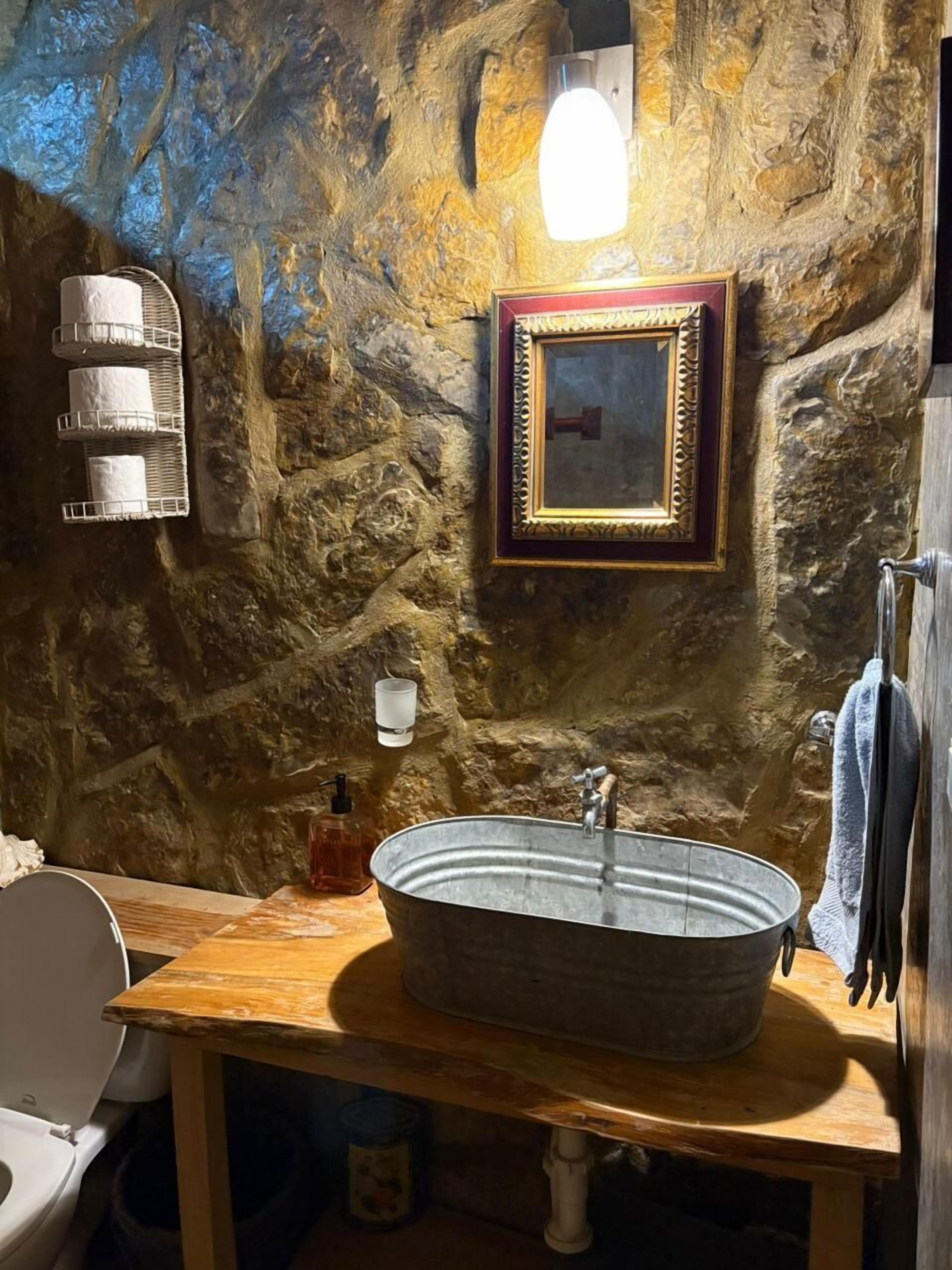 Un baño rústico con pared de piedra, tocador de madera, lavamanos de metal, espejo en marco y perchero para toallas.