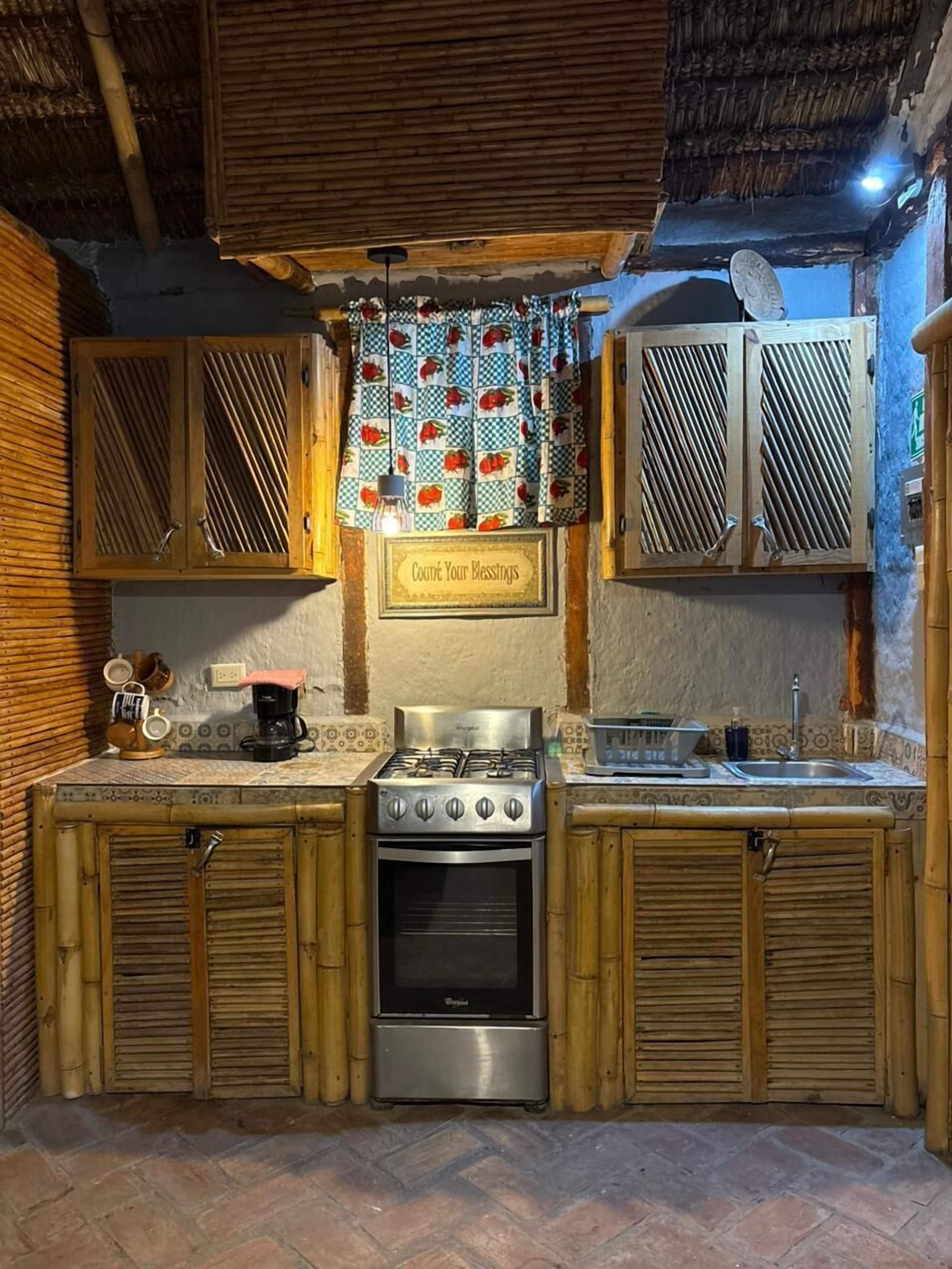 Una cocina compacta con estufa a gas, horno y lavamanos. Hay un letrero sobre la estufa que dice “Cuenta tus bendiciones”. La cocina tiene armarios de madera y techo de paja.