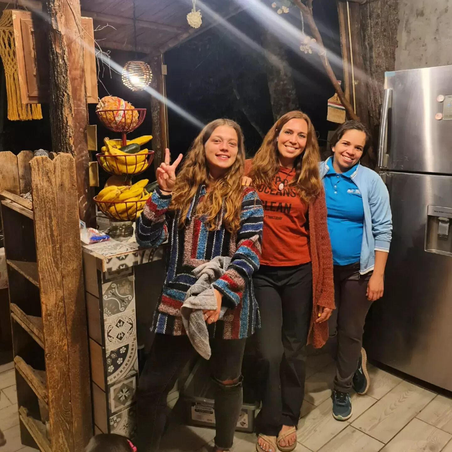 Una cocina con refrigerador, repisa de madera con plátanos y una lámpara colgante.