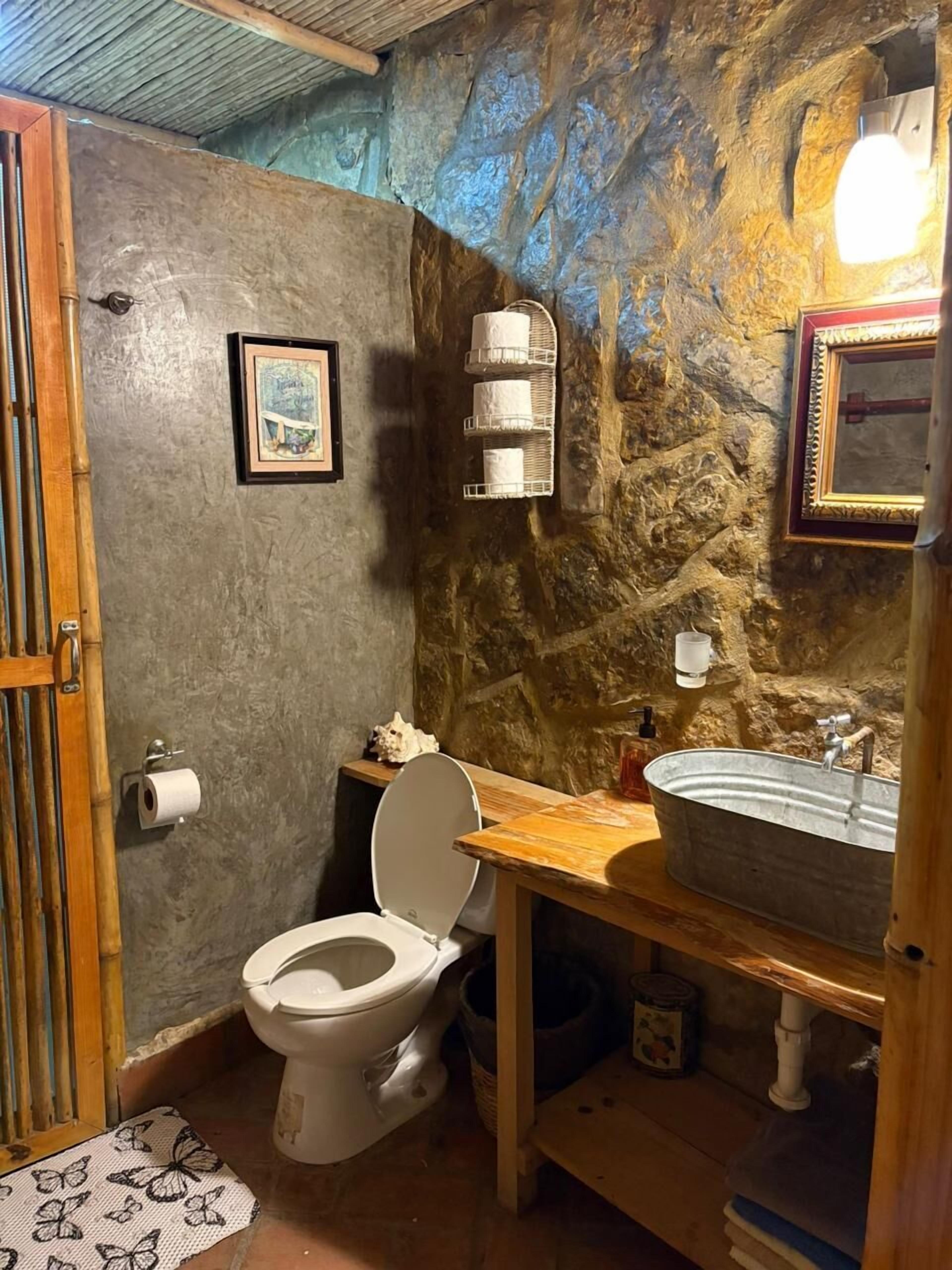 Un baño con pared de piedra rústica, tocador de madera, inodoro blanco y lavamanos de metal.
