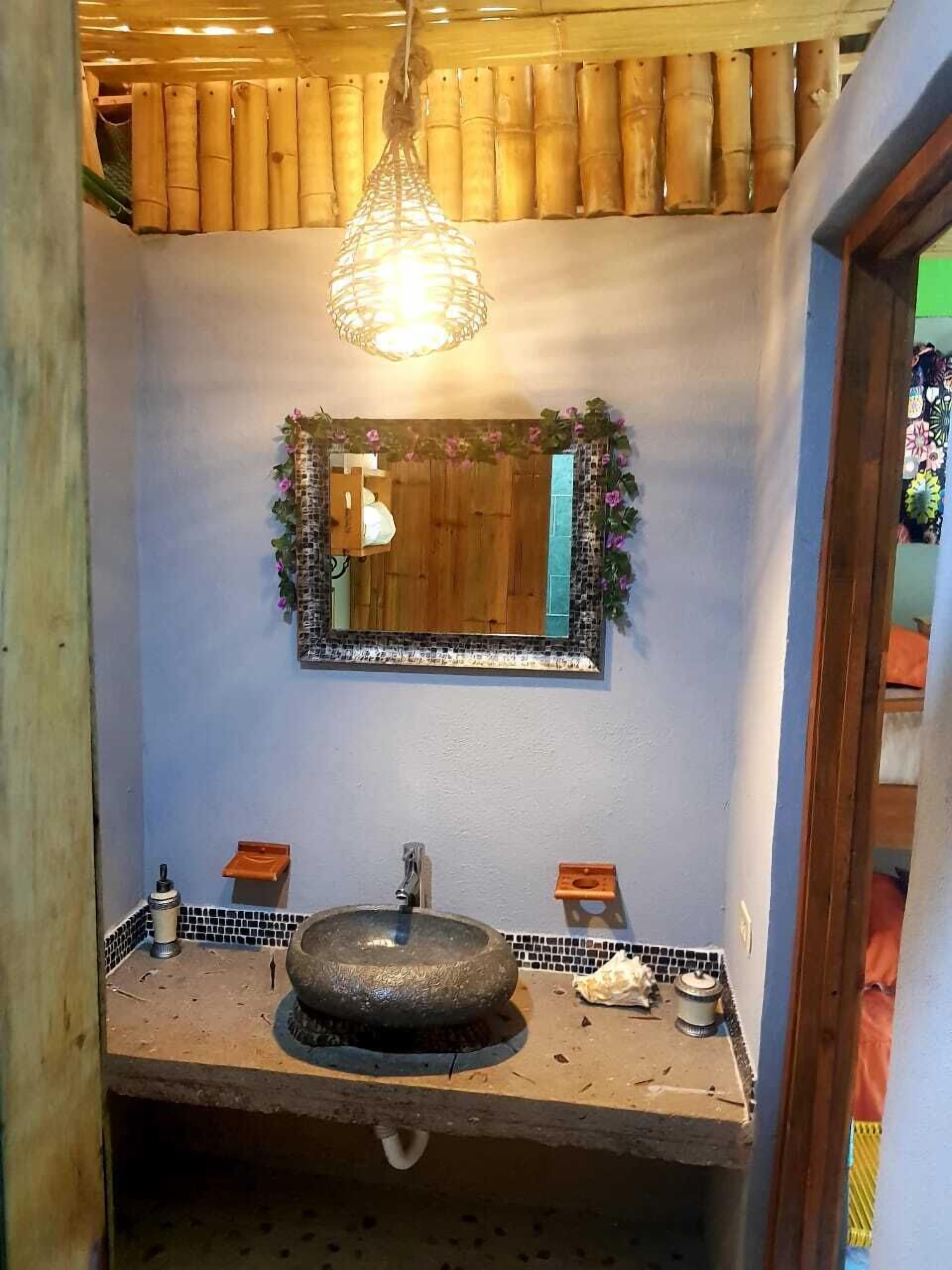 Un baño con un lavamanos de piedra, un tocador de madera, un espejo con una guirnalda floral y un aplique colgante.