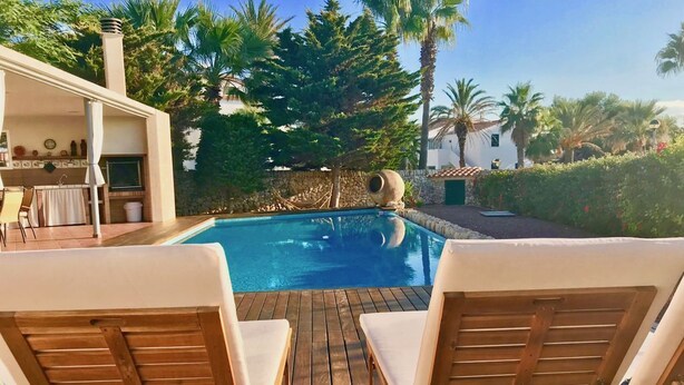 A wooden deck with two lounge chairs facing a swimming pool, surrounded by greenery and a stone wall. - VILLA VICTORIA (SON BLANC) (Son Blanc)