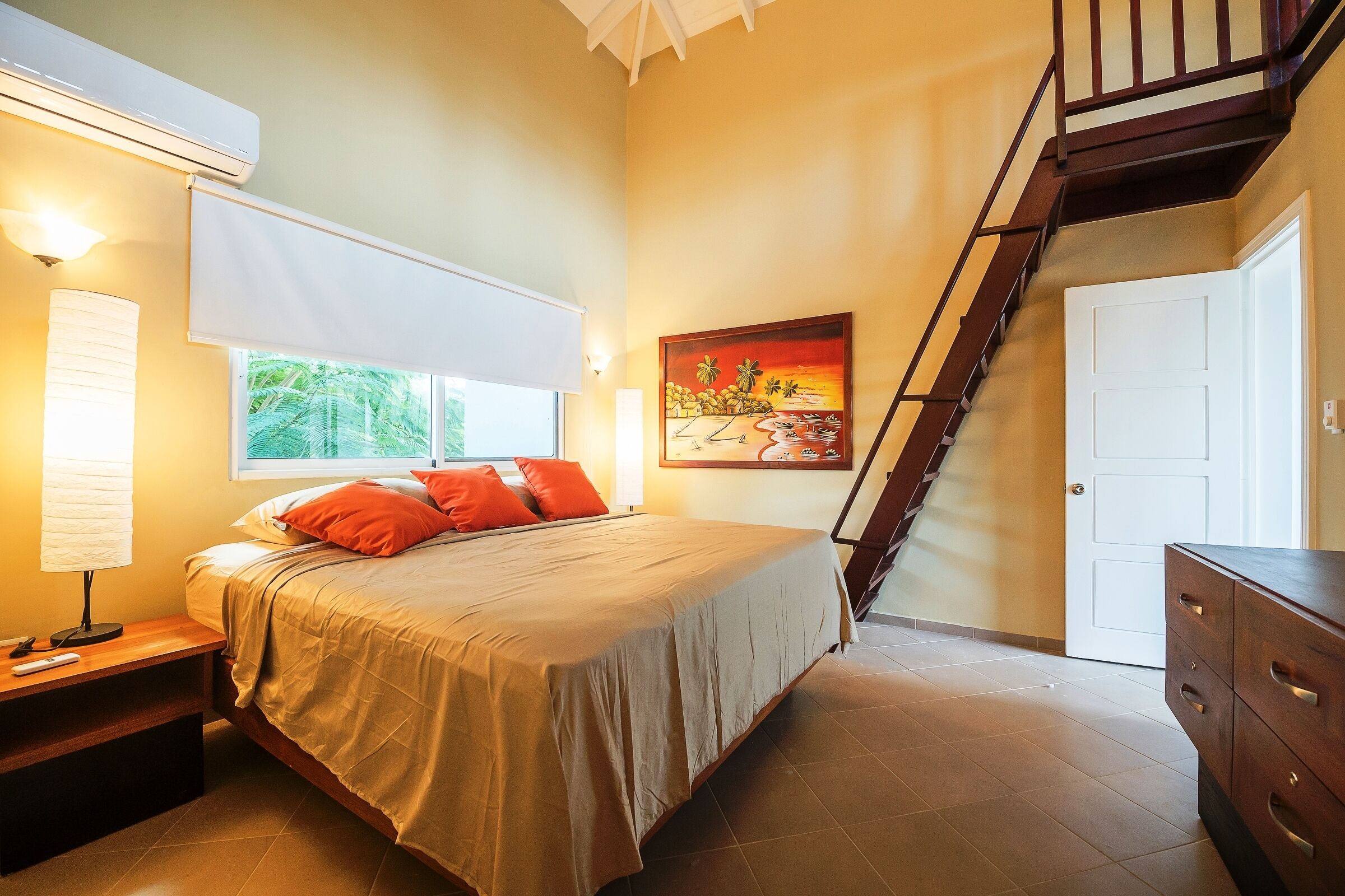 Une chambre à coucher comprenant un lit, une table de chevet, une lampe, une fenêtre avec des stores, un tableau au mur, un escalier et une porte.