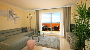 Ein Wohnzimmer mit einer Couch, einem Glastisch, einem pinkfarbenen Pouf, einer großen Topfpflanze und Blick auf eine Terrasse mit Tisch und Stühlen.