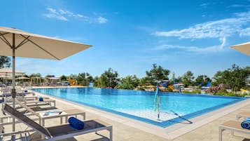 Piscina con sillones de descanso y sombrillas, rodeada de árboles y un cielo despejado.
