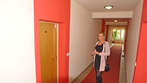 A hallway with red walls and a red carpet, a wooden door, and a person holding keys. - HoldLux Apartmanok (Sarvar)