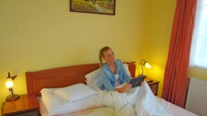 A person lying in bed with a tablet, in a room with yellow walls, a wooden headboard, and two bedside lamps. - HoldLux Apartmanok (Sarvar)
