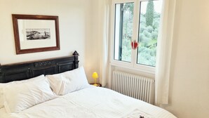 A neatly made bed with white linens, a framed picture on the wall, a window with a view of greenery, and a bedside table with a lamp.