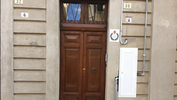 Une porte à deux battants en bois, dotée d’une poignée en métal, flanquée de murs recouverts d’un revêtement beige et percés de fenêtres numérotées.