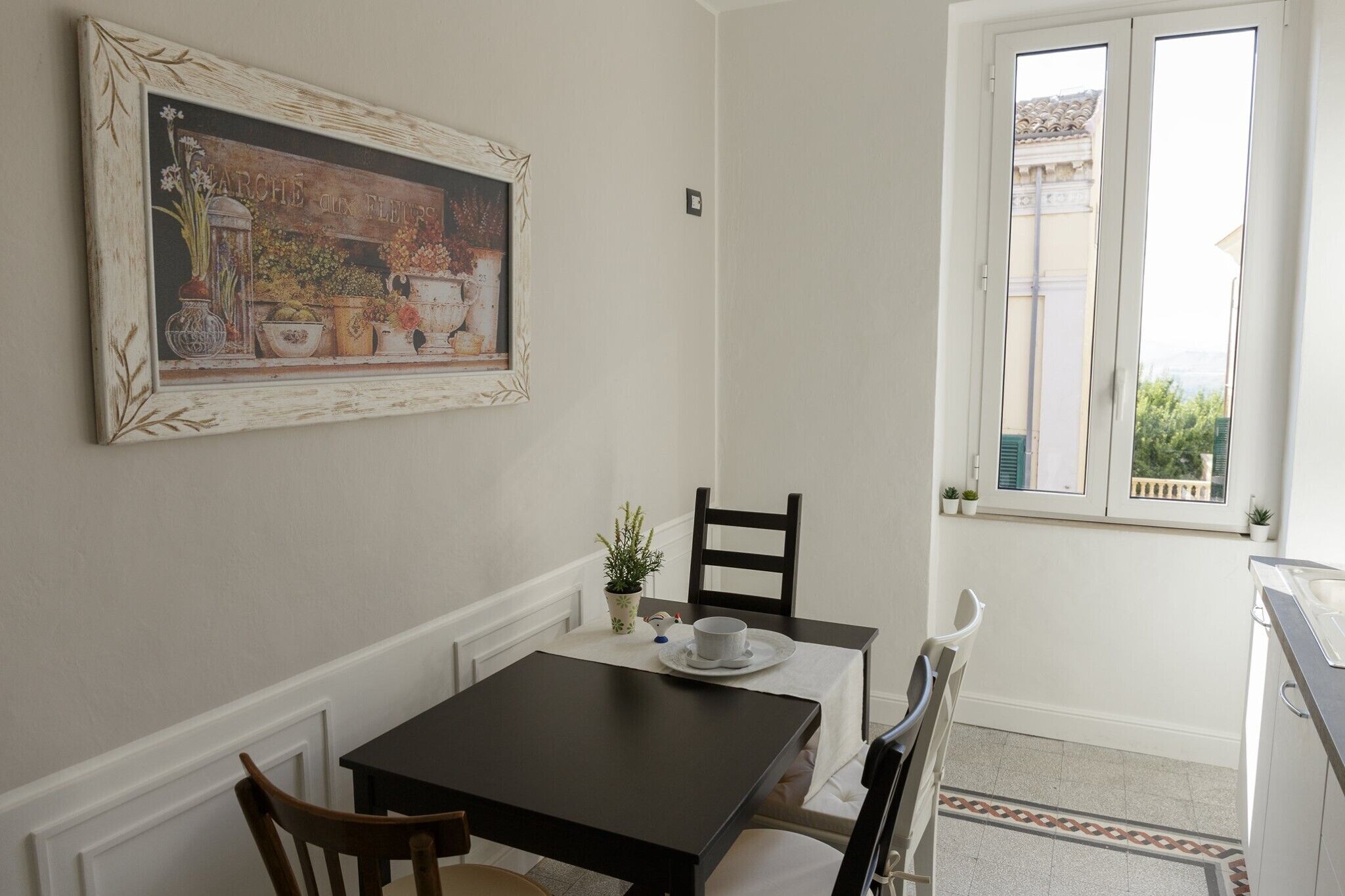 A dining area with a table, chairs, a painting, a window, and a small potted plant.