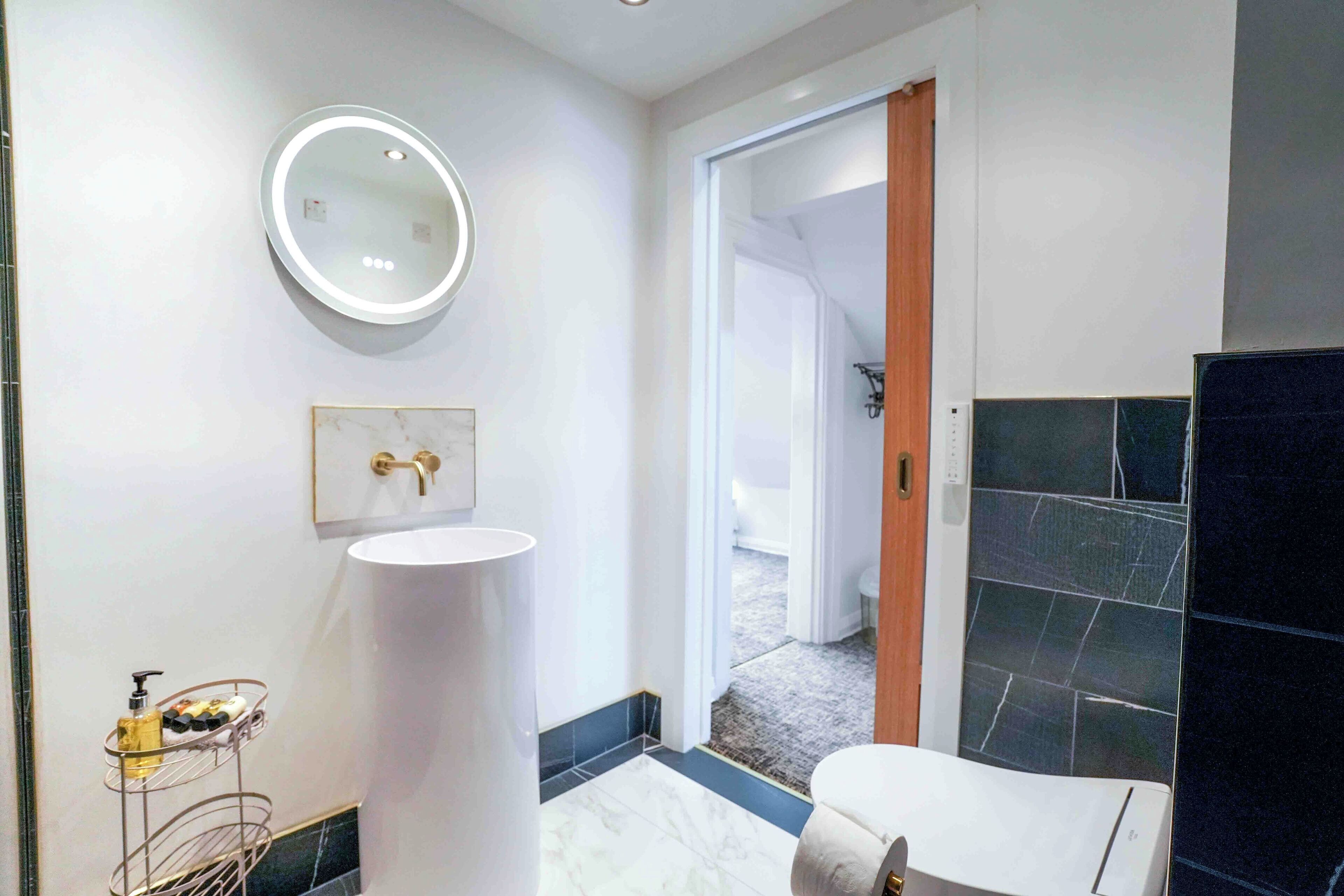 Un baño moderno con un espejo redondo, lavamanos blanco y zona de ducha con azulejos oscuros.