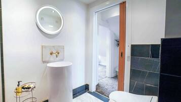 Un baño moderno con un espejo redondo, lavamanos blanco y zona de ducha con azulejos oscuros.
