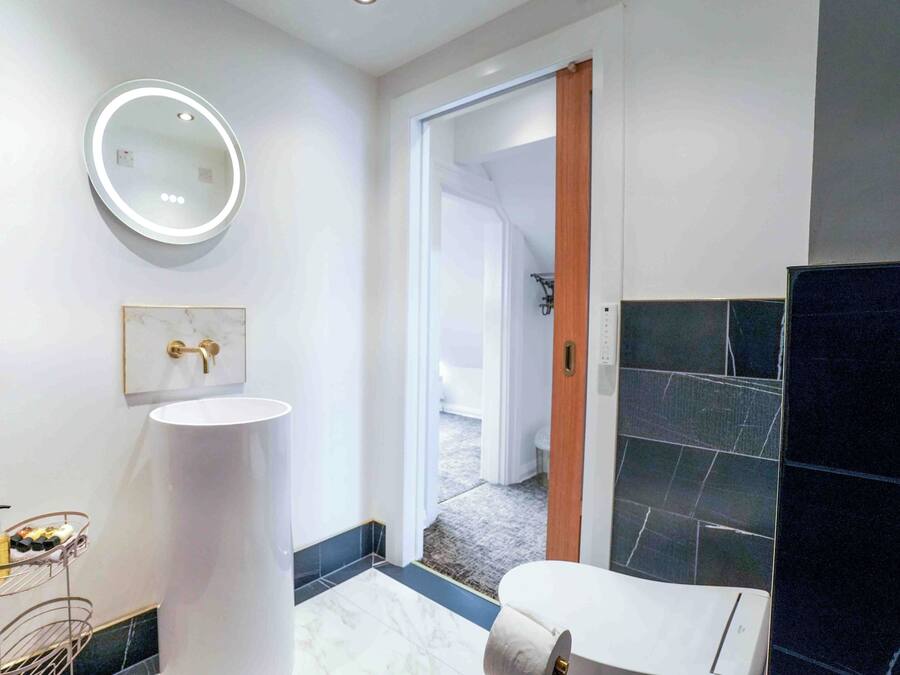 Un baño moderno con un espejo redondo, lavamanos blanco y zona de ducha con azulejos oscuros.