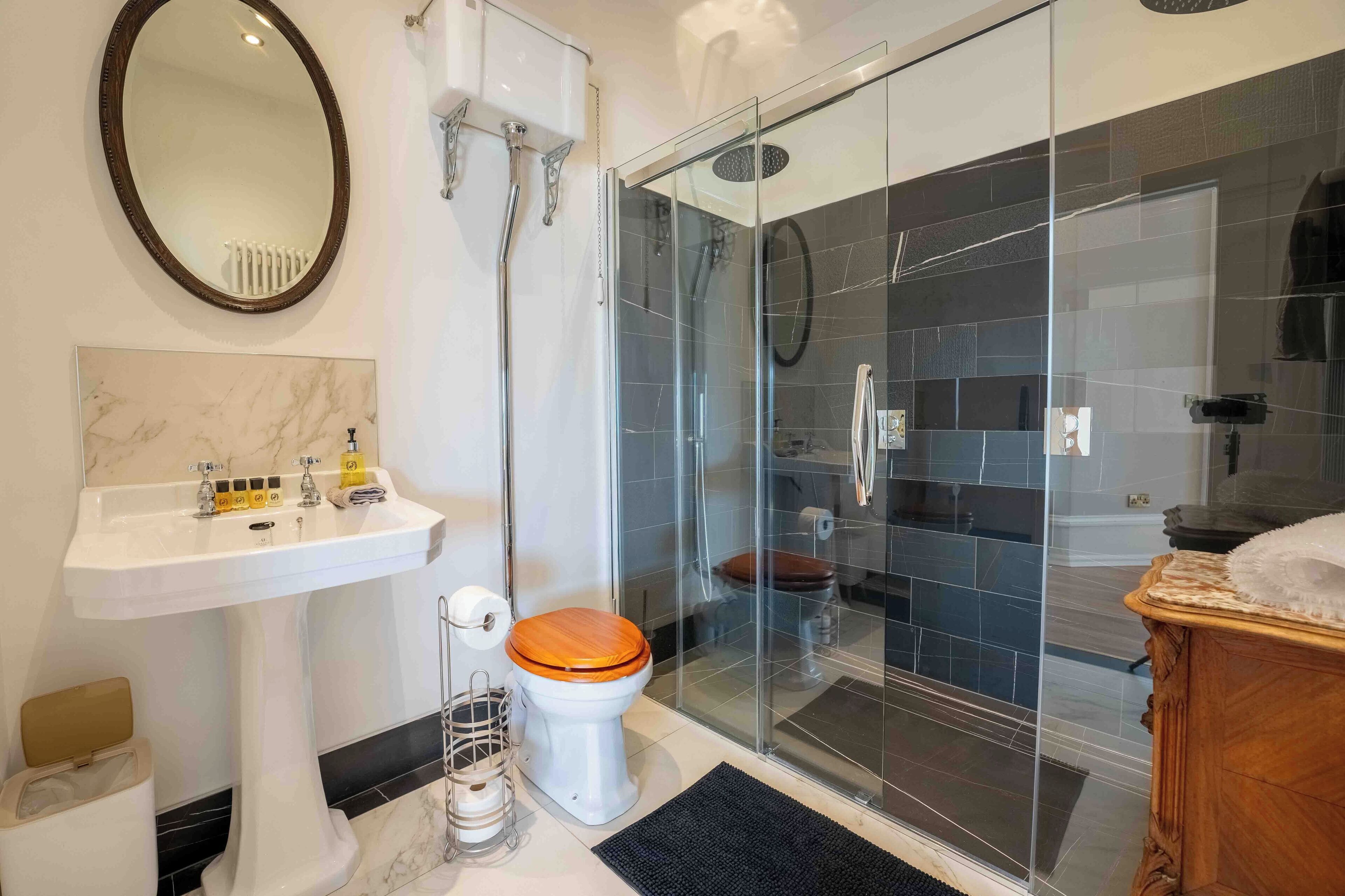 A modern bathroom with a glass-enclosed shower, a white pedestal sink, and a wooden toilet.