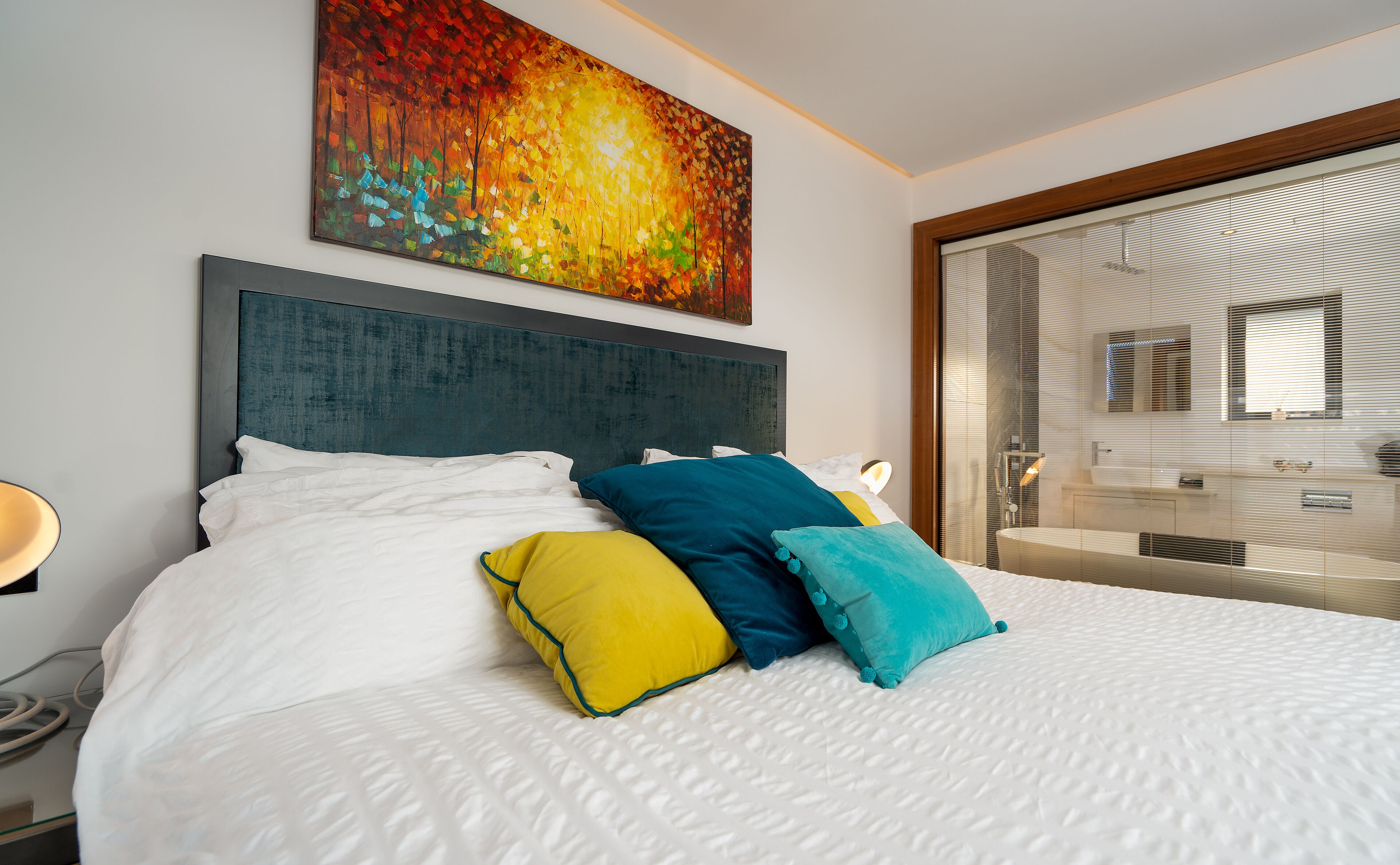 A modern hotel room with a large bed, a headboard, and a colorful painting above it.
