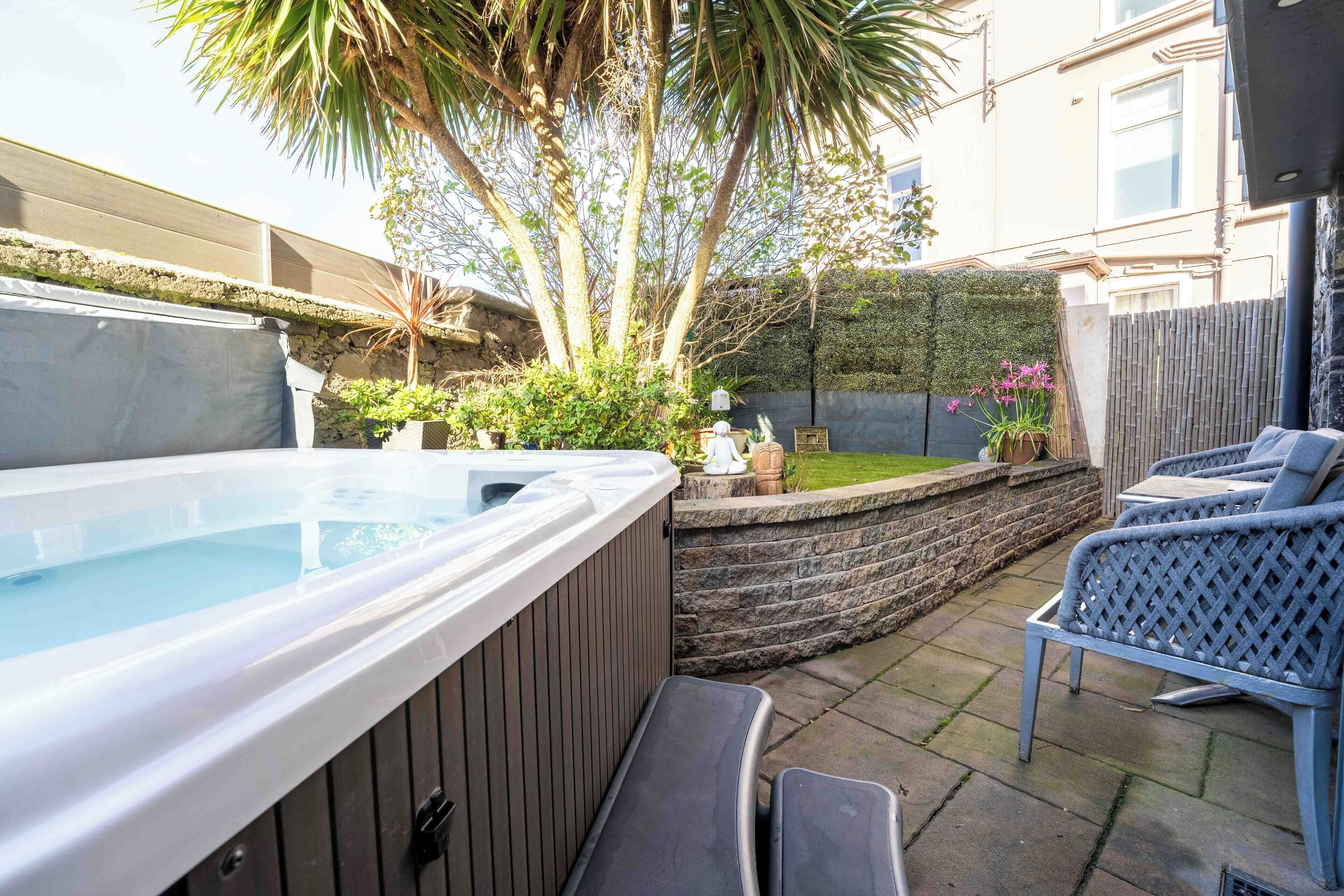 A hot tub with a wooden exterior, a stone wall, and a seating area with wicker chairs.