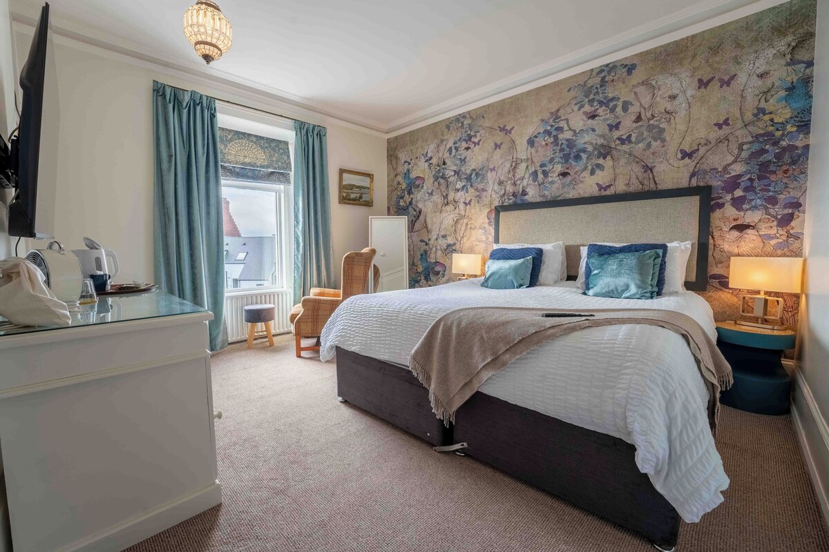 A hotel room with a large bed, a desk with a telephone, a chair, a window with curtains, and a floral wallpaper.