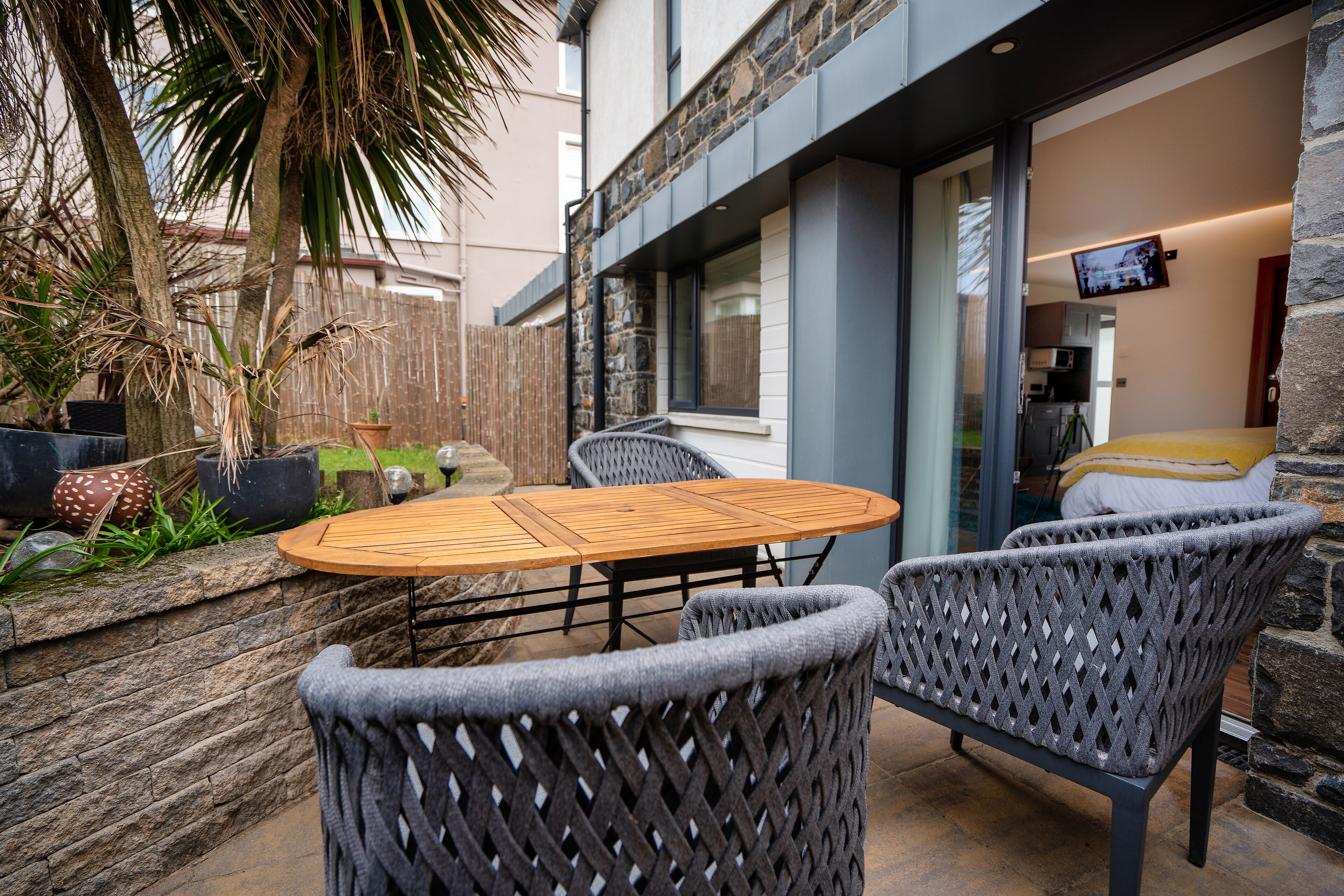 A patio area with a round wooden table and two wicker chairs, a stone wall, and a view into the building.