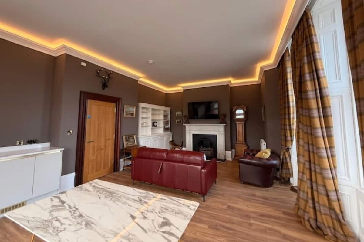 Un salon avec une cheminée, un canapé en cuir rouge, une table basse en marbre et un rideau à carreaux.