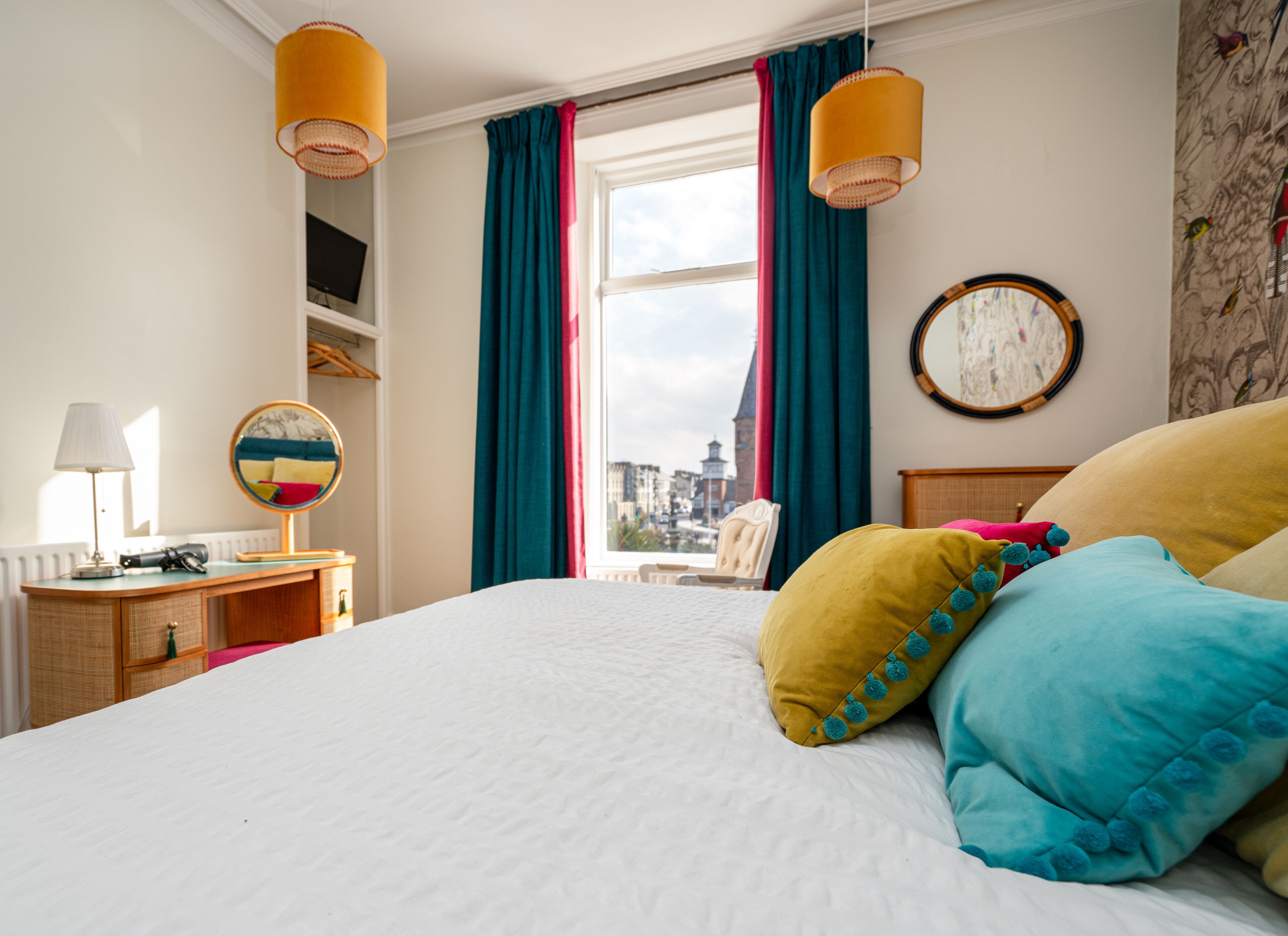 A hotel room with a large bed, a desk with a lamp, a chair, and a view of the city through a window with curtains.