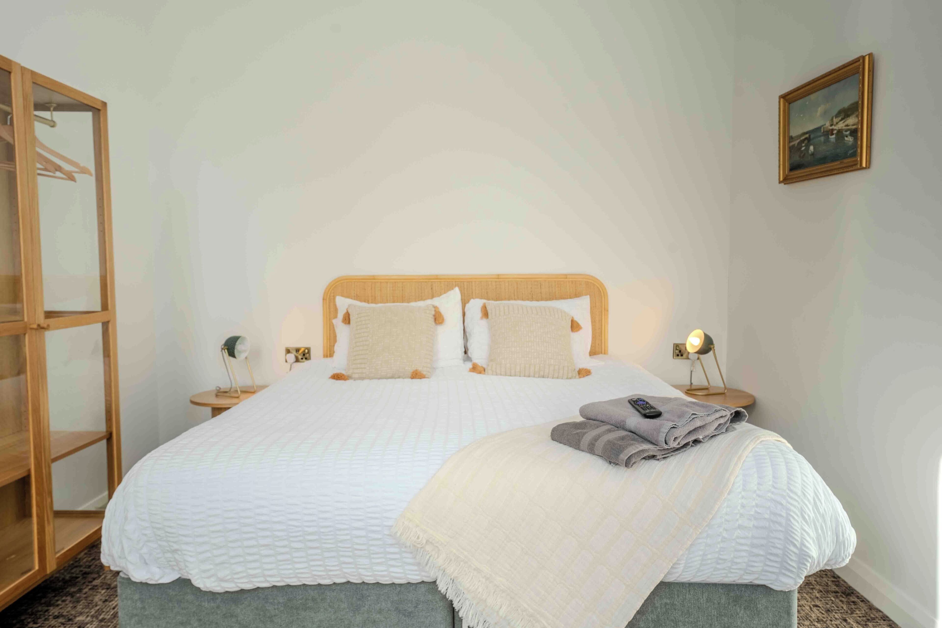 A neatly made bed with a white quilt, two pillows, and a folded blanket on the side. There is a bedside table with a lamp and a small decorative item.