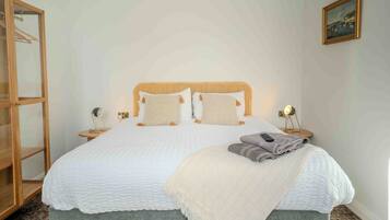 A neatly made bed with a white quilt, two pillows, and a folded blanket on the side. There is a bedside table with a lamp and a small decorative item.
