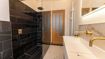 Een moderne badkamer met een douchegedeelte betegeld in zwart, een houten deur, een witte wastafel met gouden details en een marmeren spatwand.