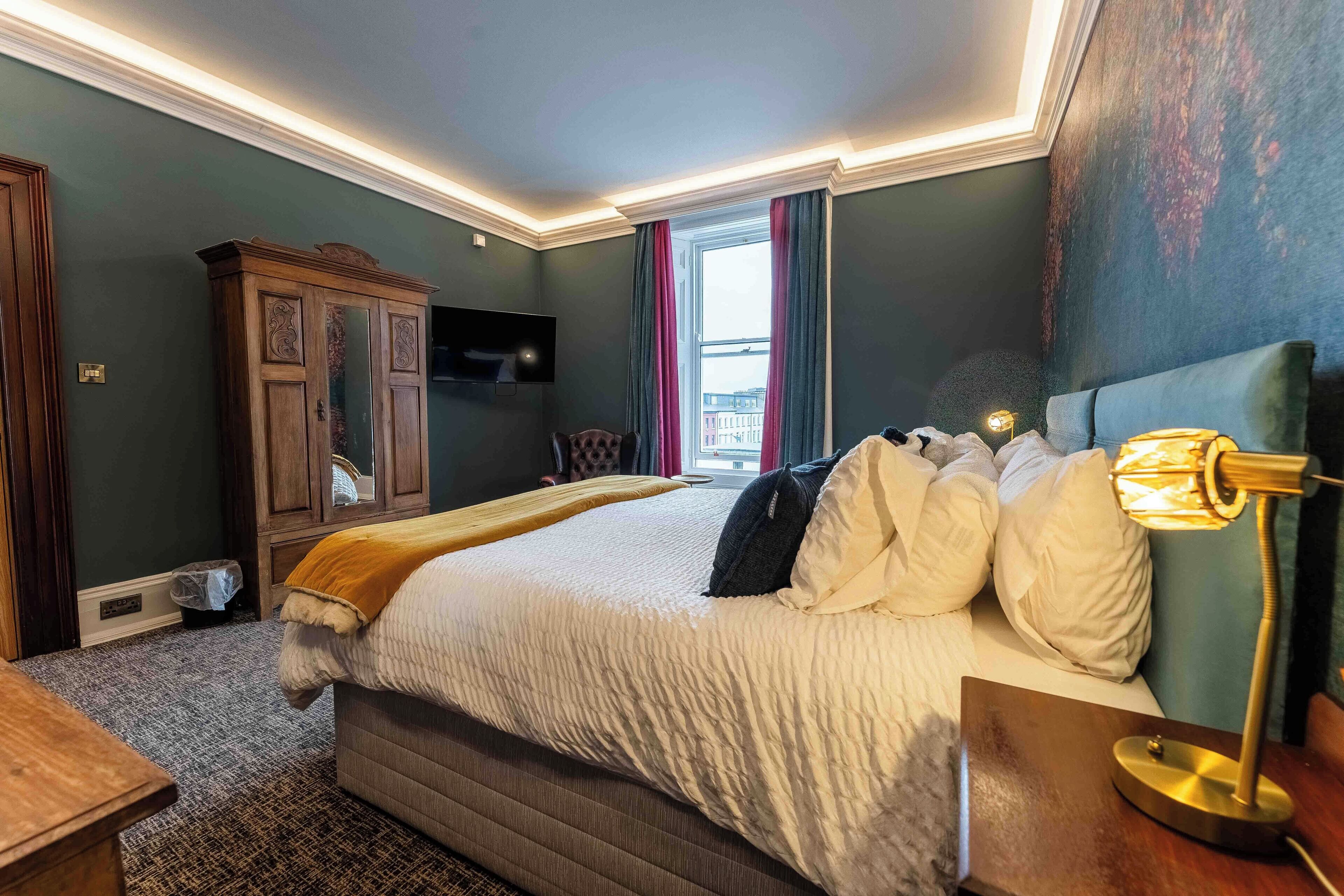 A neatly arranged bedroom with a large bed, a wooden cabinet, a flat-screen TV, and a window with curtains.
