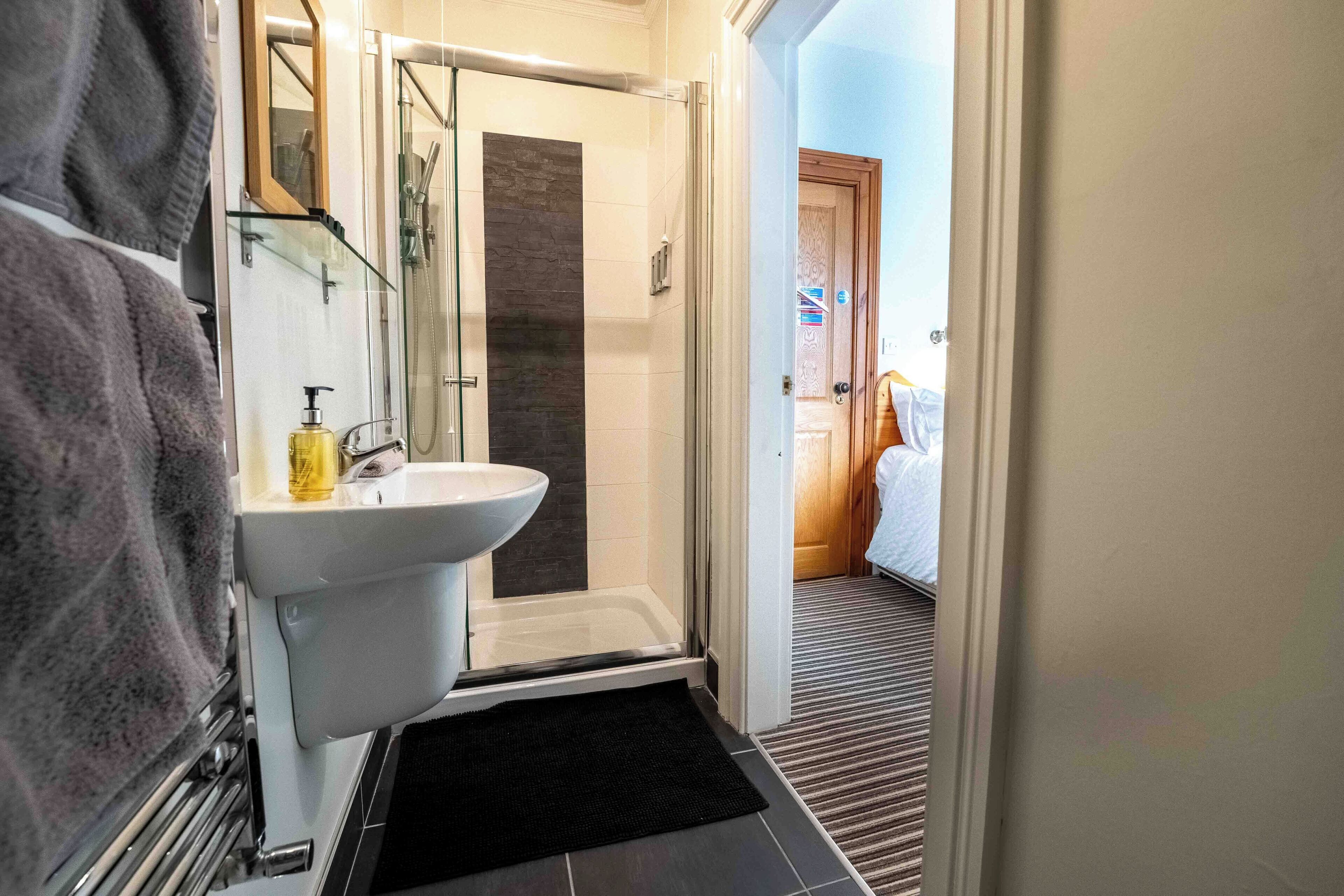 Une salle de bain moderne avec une douche à l’italienne en verre, un lavabo blanc et un tapis noir menant à une chambre.