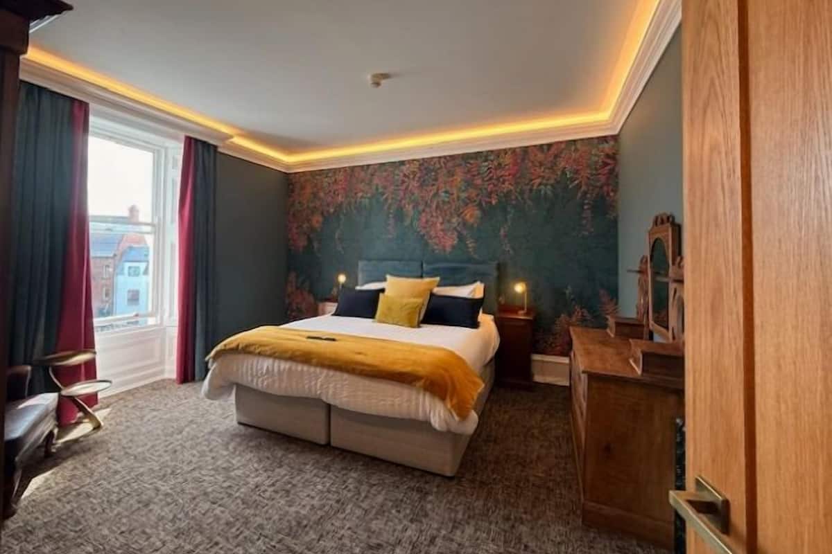 Une chambre d’hôtel avec un grand lit, une table de chevet, une commode en bois et une vue sur les bâtiments à travers la fenêtre.
