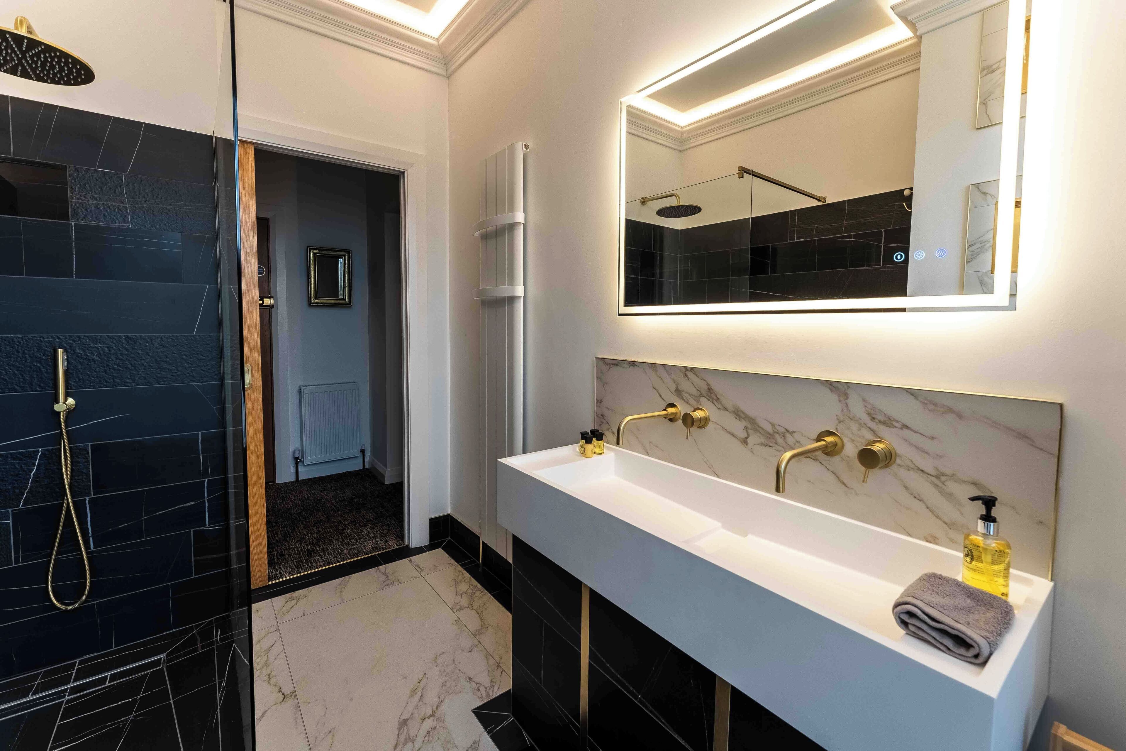 Een moderne badkamer met een douchecabine van glas, een wit badkamermeubel met een marmeren spatwand en een grote spiegel boven de wastafel.