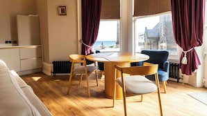 A cozy room with a round wooden table, two chairs, a blue sofa, and a view of the sea through large windows with curtains. - Antrim House Guest Rooms & Suites (Portrush)