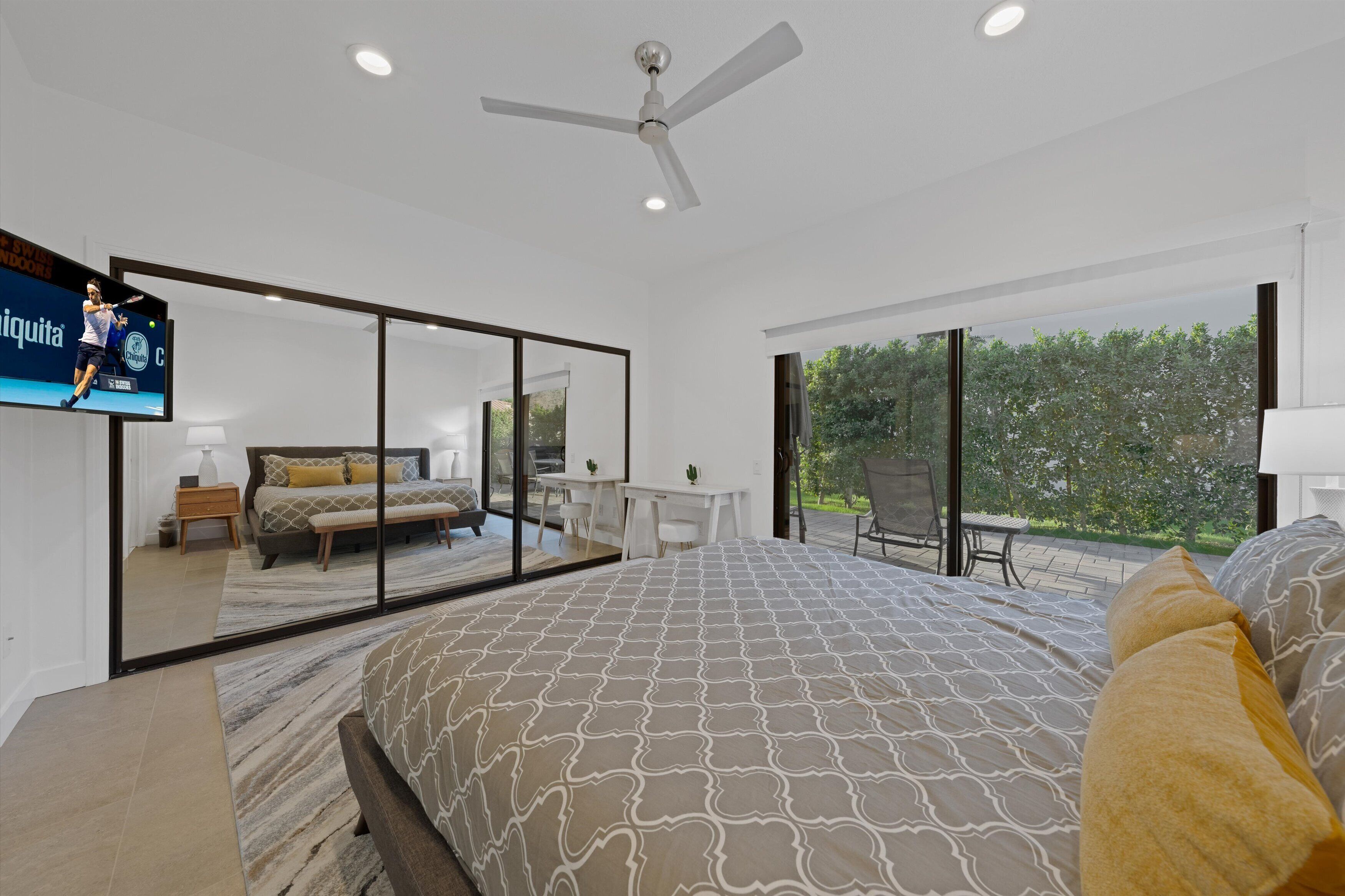 A modern bedroom with a large bed, a ceiling fan, a TV, and a balcony with a view of greenery.
