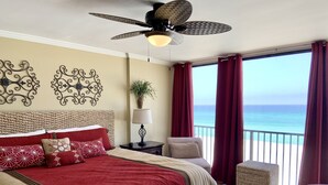 A hotel room with a large bed, a ceiling fan, a nightstand with a lamp, a chair, and a view of the ocean.