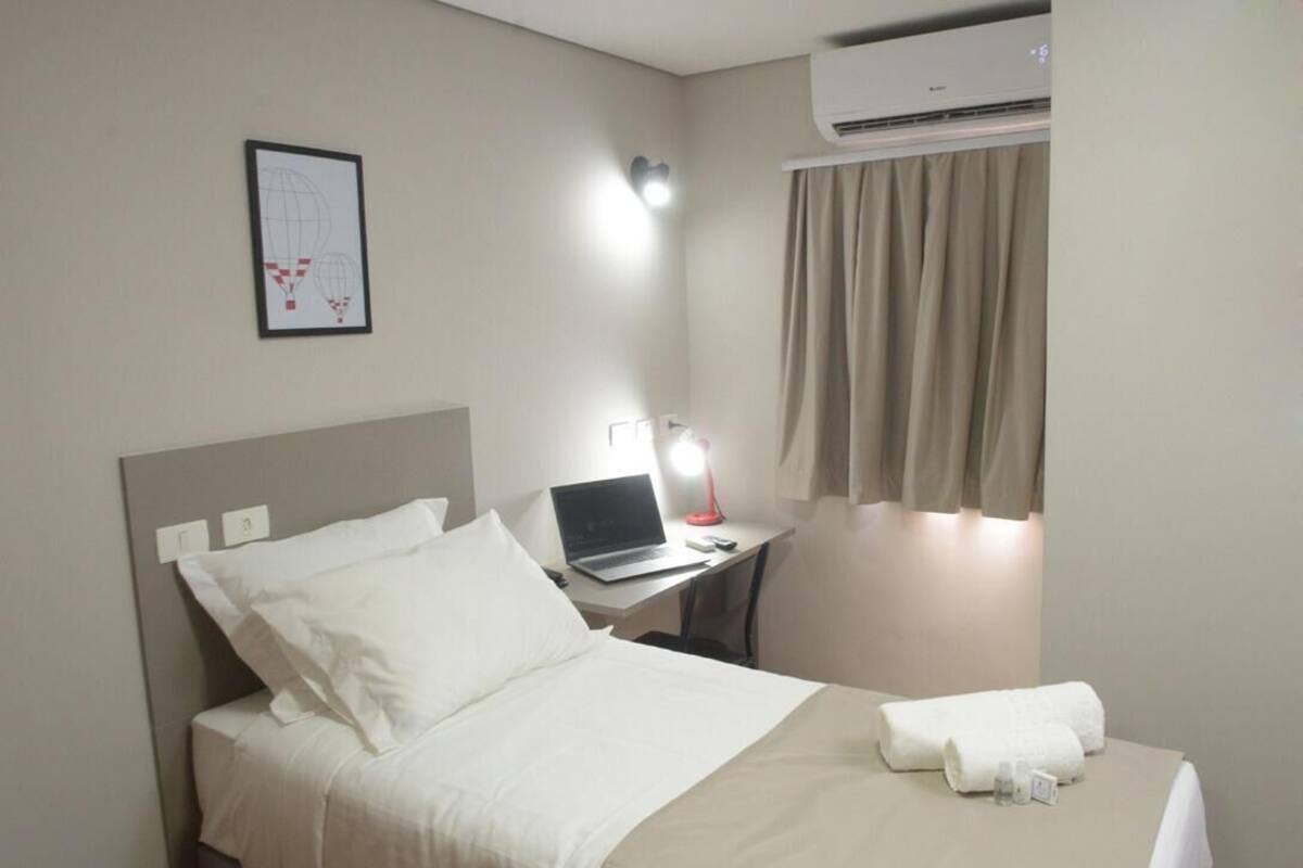 A hotel room with a bed, a desk with a laptop, a desk lamp, an air conditioner, and a framed picture on the wall.