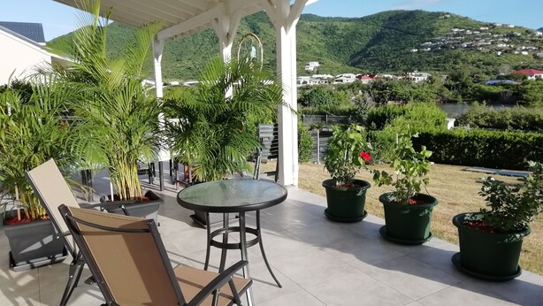 Een overdekt terras met een tafel en stoelen, omgeven door planten in potten en met uitzicht op een mooi landschap van heuvels en huizen.