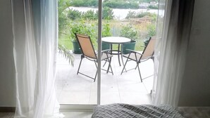 Een kamer met een bed, uitzicht op een balkon met stoelen en een tafel, en een groot raam met dunne gordijnen.