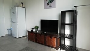 Een woonkamer met een witte koelkast, een flatscreen televisie op een houten standaard, rieten manden en een zwart kastenrek.