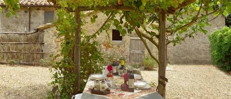 A table set up under a pergola with a stone wall and rustic building in the background.