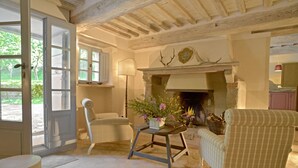 Une pièce chaleureuse avec une cheminée, un fauteuil, une table ornée de fleurs et une vue sur l’extérieur.