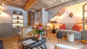 Un salon chaleureux de style loft, avec des poutres en bois apparentes, un canapé gris agrémenté de coussins rouges et blancs, une table ronde sur laquelle est posé un vase de fleurs, et une bibliothèque décorative.